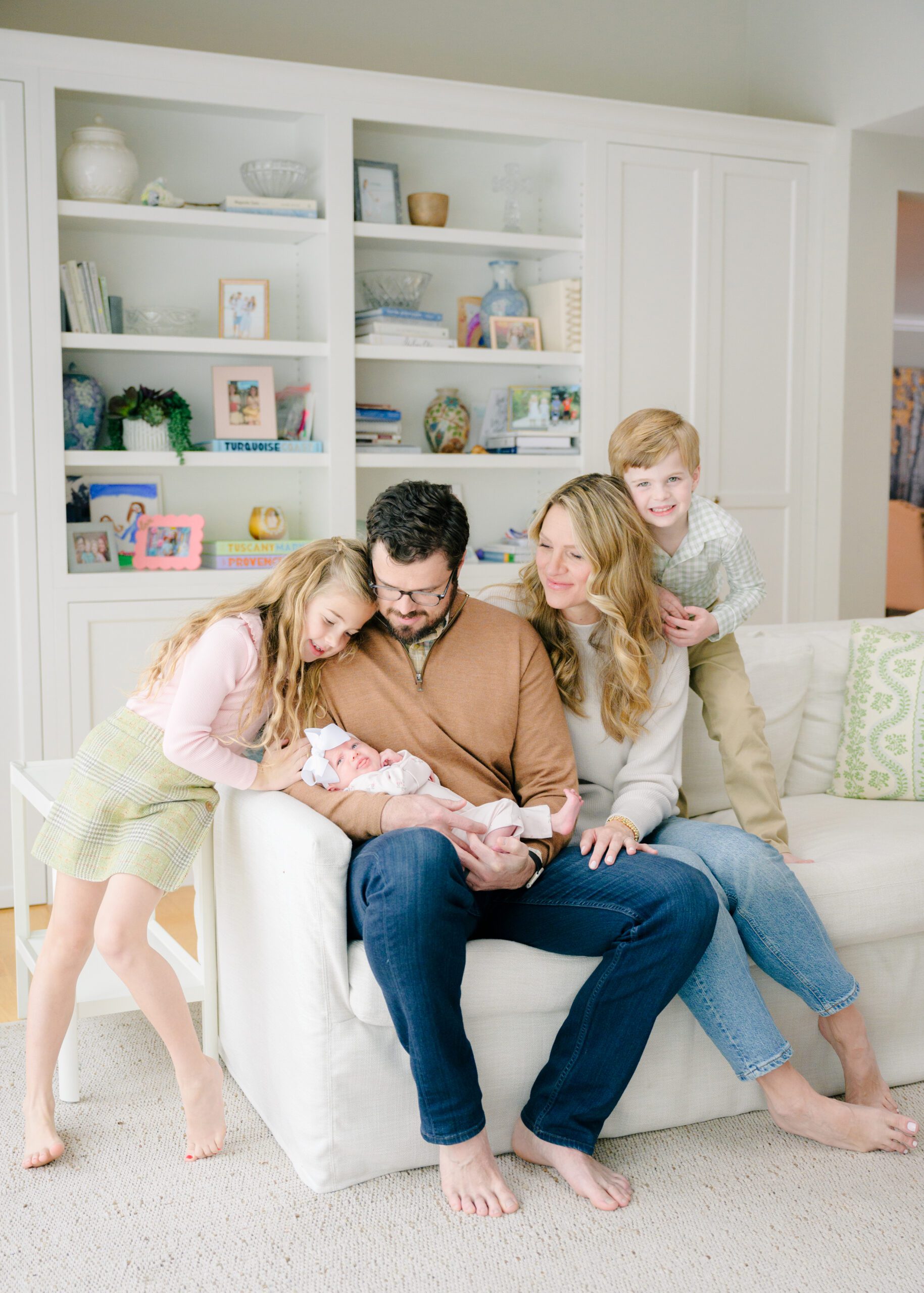 Family of five snuggled on a couch looking at a newborn baby smiling.