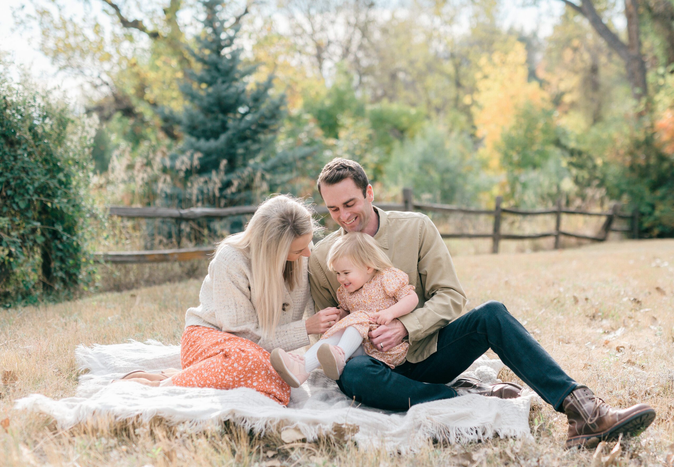 A family sitting on a blanket in a pile of leaves in the fall smiling at one another while the mom tickles her little girl.