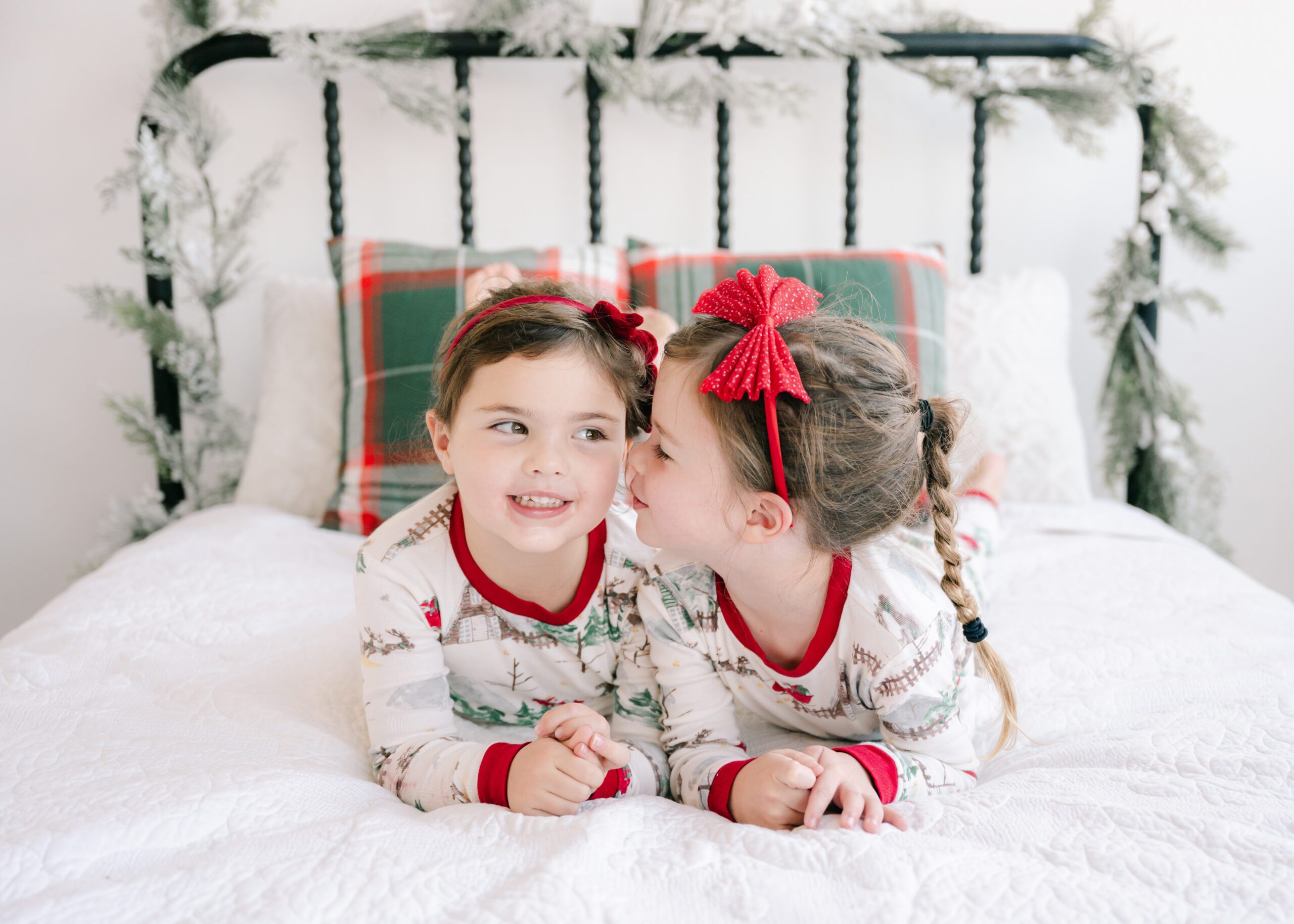 Two little girls dressed in matching christmas pjs giggling on a decorated bed.