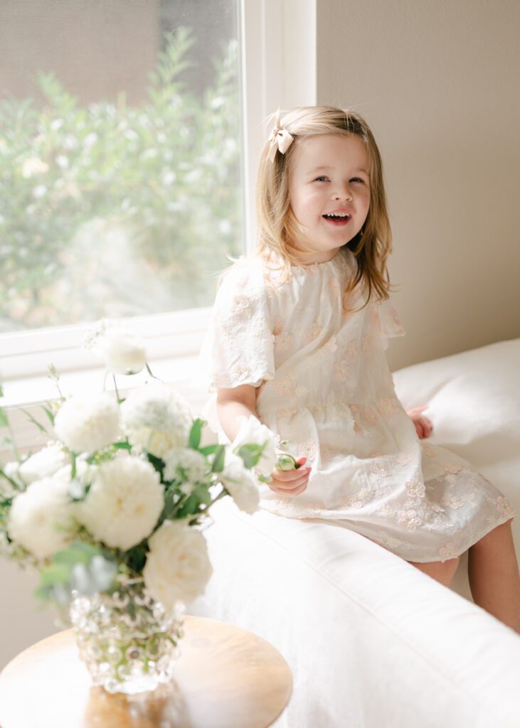 Little girl smiling up at sitting on the edge of a couch in front of a vase of fresh white flowers. Photo by Denver photographer Maegan R Photography