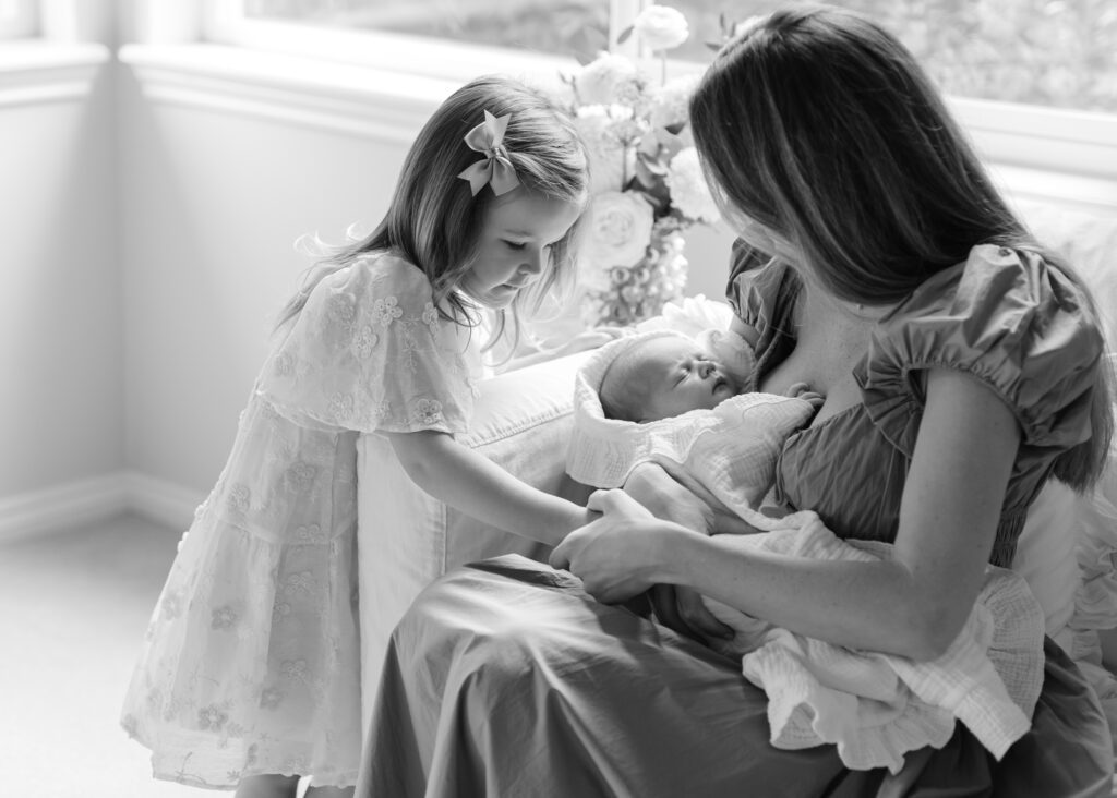 Black and white image of a mom holding her little girls hand while they look down at a newborn baby. Photo by Denver photographer Maegan R Photography