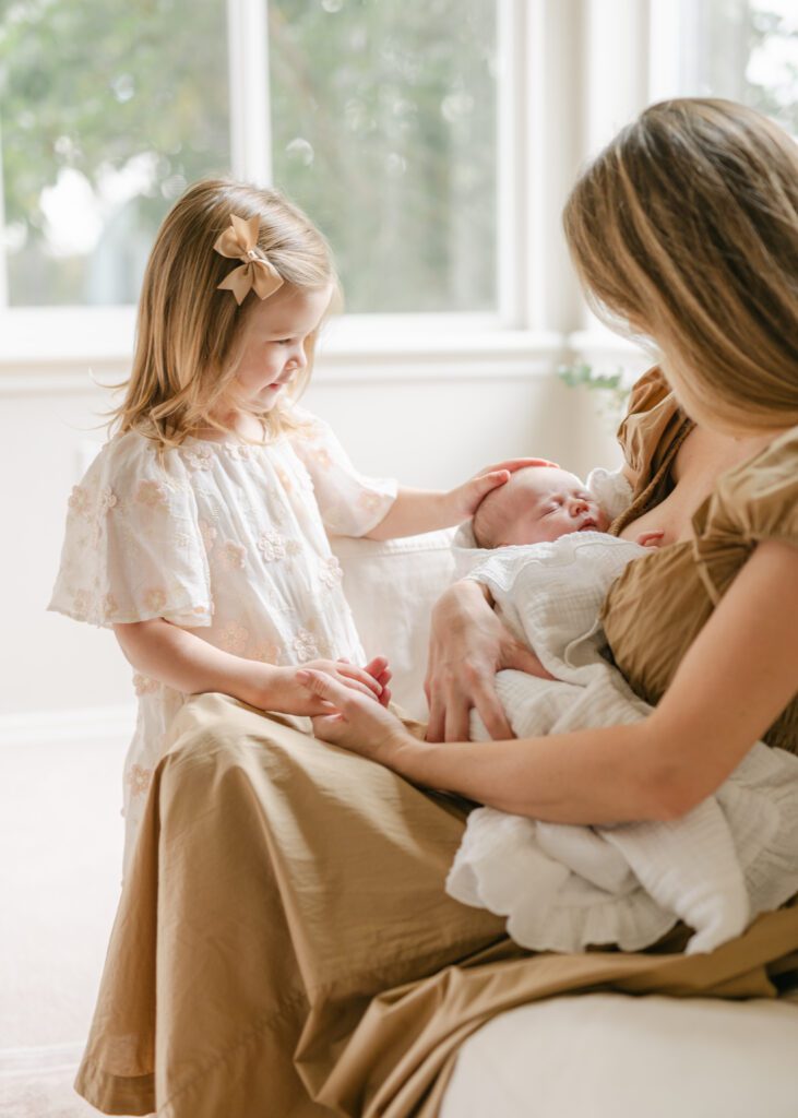 Mom holding a newborn baby in a fresh white blanket while her toddler daughter places her hand on the baby's head. Photo by Denver photographer Maegan R Photography