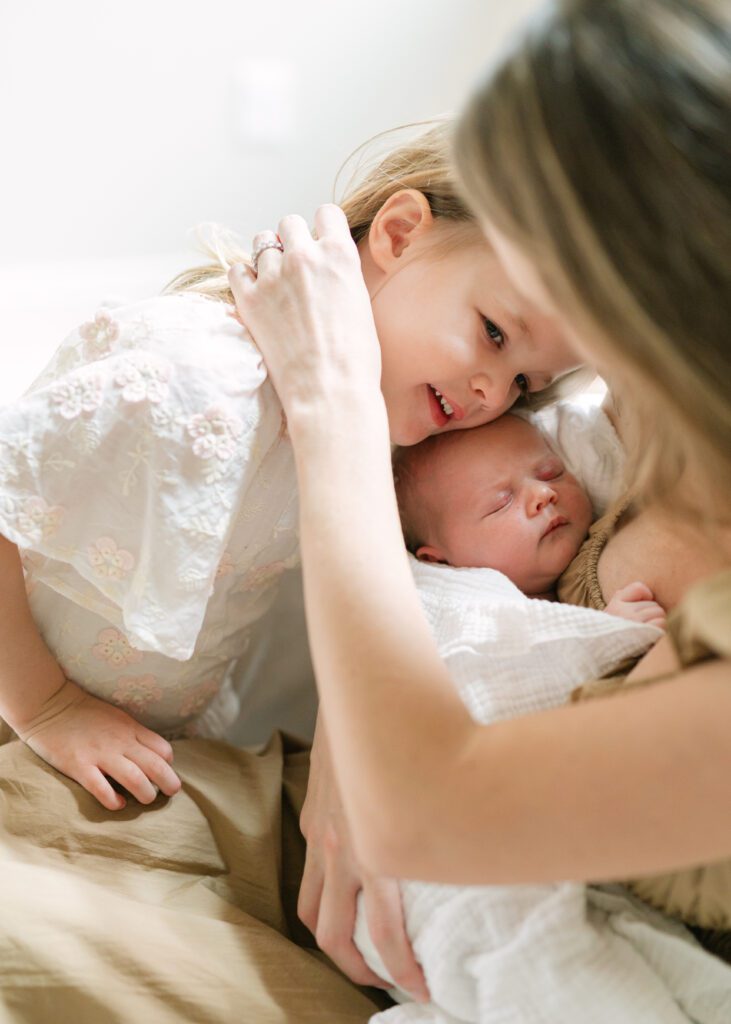 Toddler girl hugging a newborn baby while her mom gently moves her hair behind her ear. Photo by Denver photographer Maegan R Photography