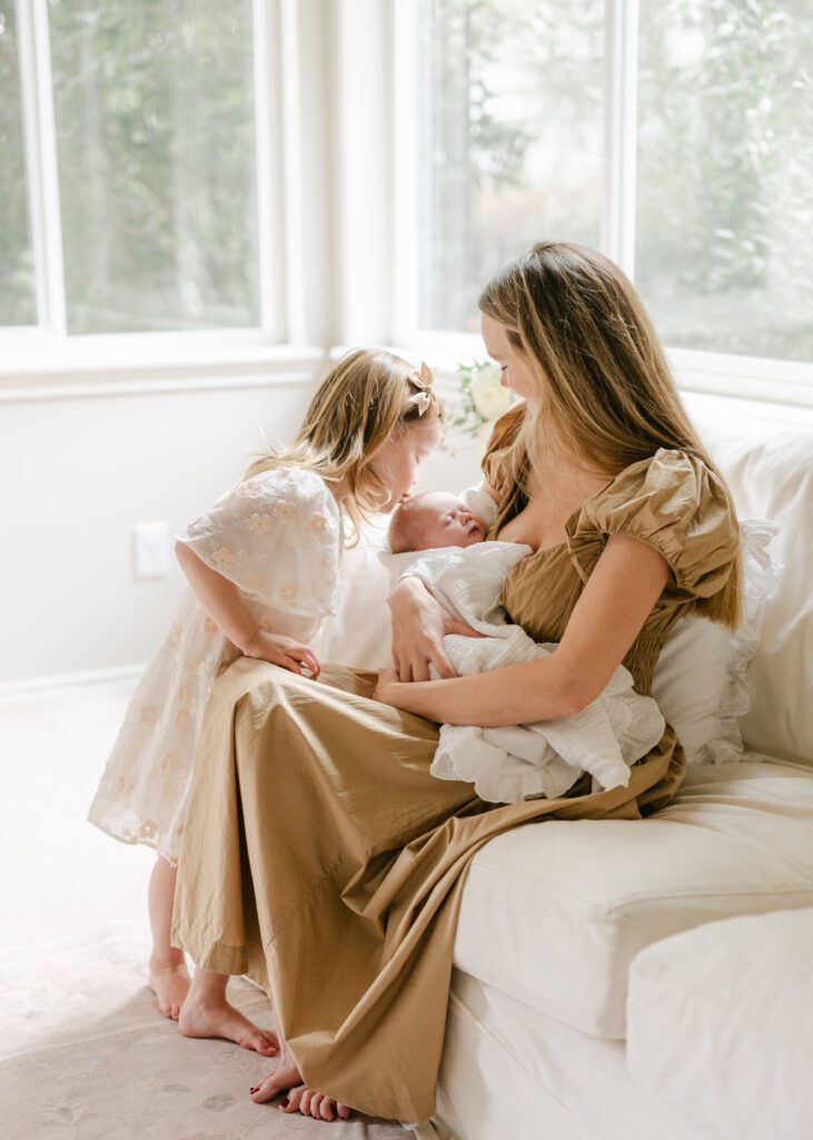 Mom holding a newborn baby in a fresh white blanket with the older sister kisses the baby's forehead. Photo by Denver photographer Maegan R Photography