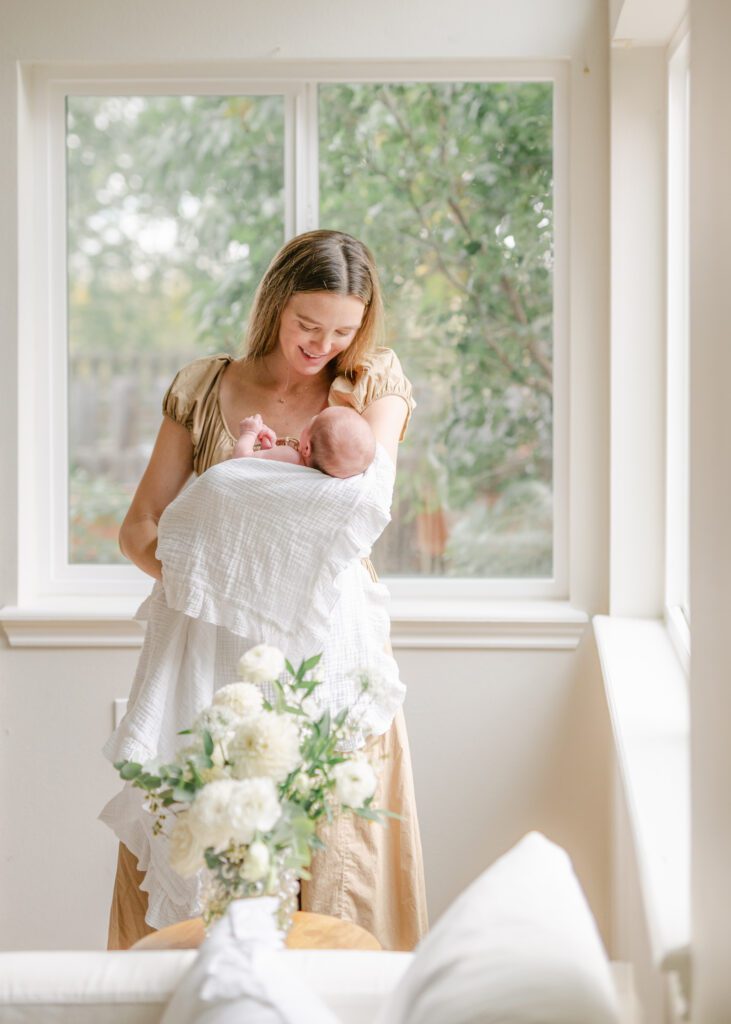 Pulled back image of a mom in an elegant tan dress holding her tiny baby in a fresh white swaddling blanket with a vase of white flowers in front of the. Photo by Denver photographer Maegan R Photography