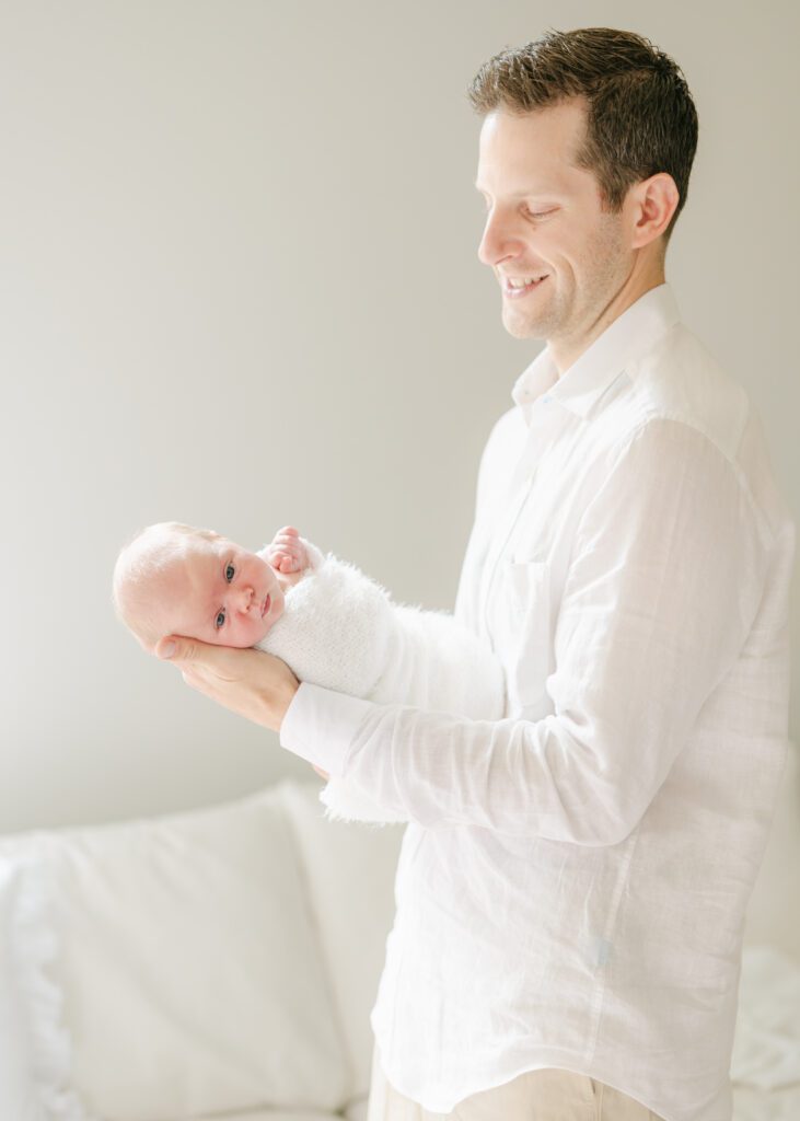 Dad in a classic white button down holding his swaddled newborn baby. Photo by Denver photographer Maegan R Photography