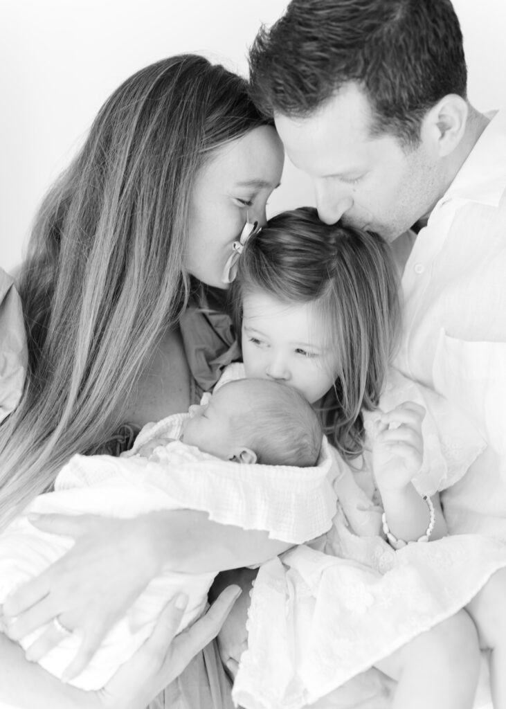 Black and white image of a family of four.  The mom and dad are kissing their toddler daughter's head while she kisses a newborn baby.  Photograph by Maegan R Photography. 