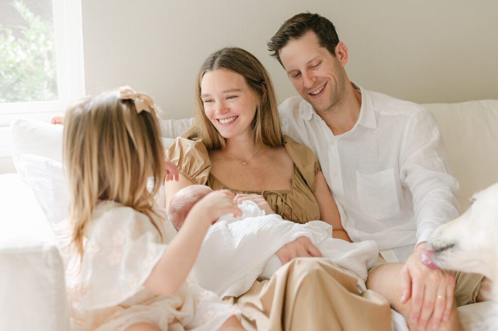 Mom and dad sweetly smiling at her toddler daughter while holding her newborn baby. Photograph by Maegan R Photography. 