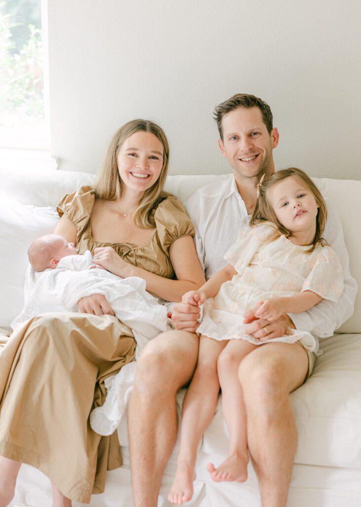 Family of four sitting in a white couch, dressed elegantly in white and tan celebrating the new addition to their family. Photograph by Maegan R Photography. 