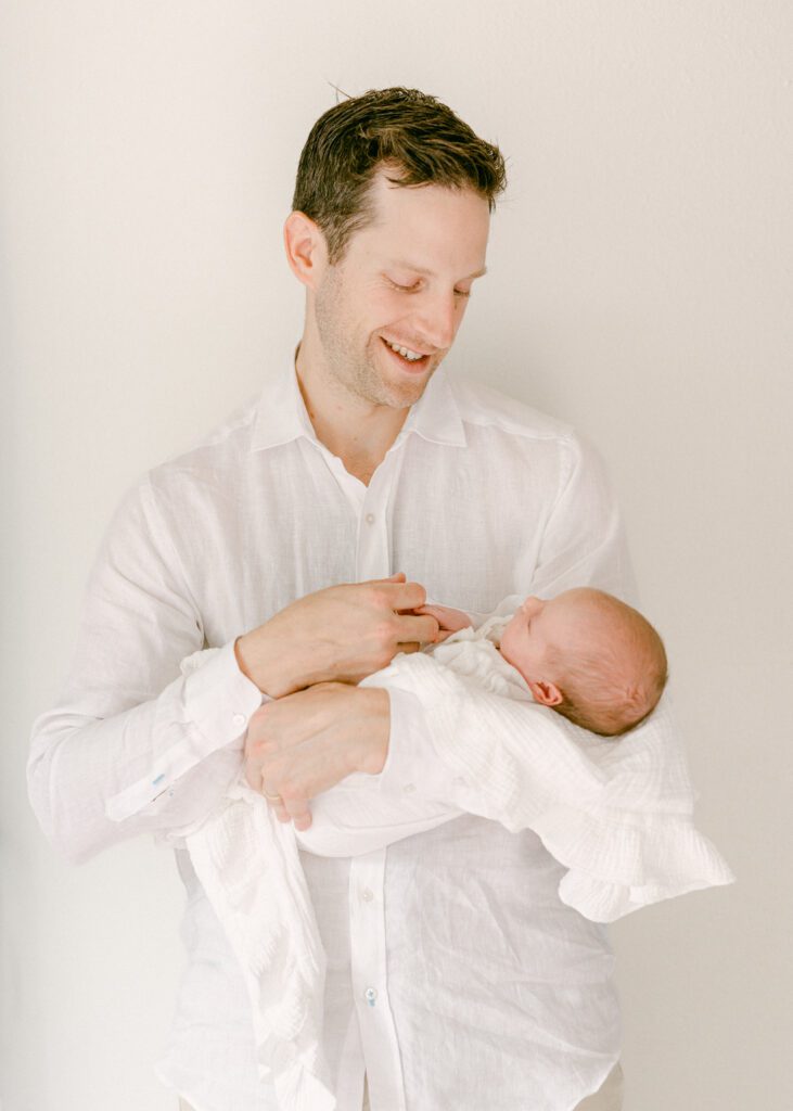 Dad craddling his newborn baby smiling down at her while he holds the baby's hand. Photograph by Maegan R Photography. 