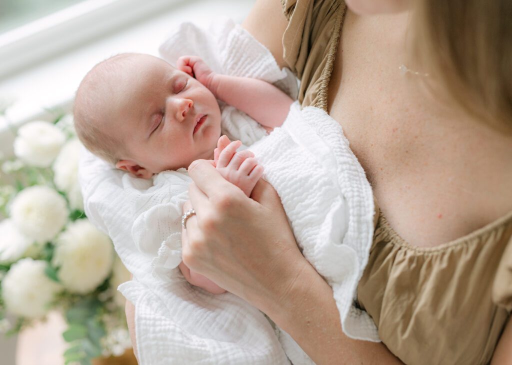 Close up image of a newborn baby sleeping in a ruffled white swaddle blanket with a woman holds the baby's hand. Photograph by Maegan R Photography. 