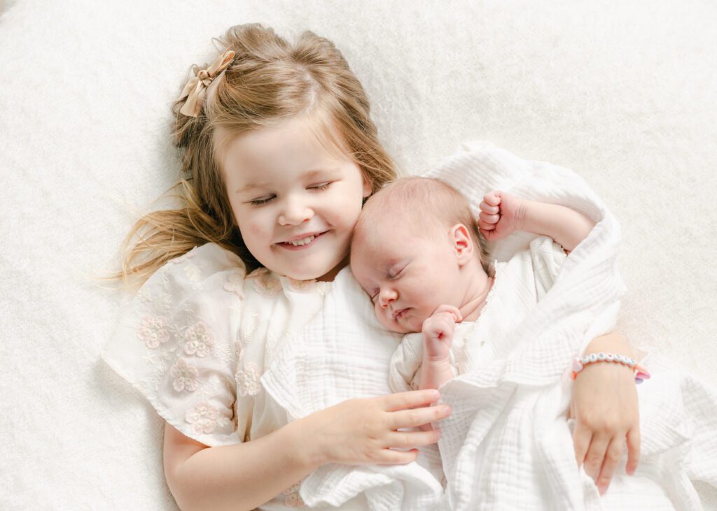 Photo of a toddler and baby snuggling laying down with their eyes closed on a white blanket. Photograph by Maegan R Photography. 