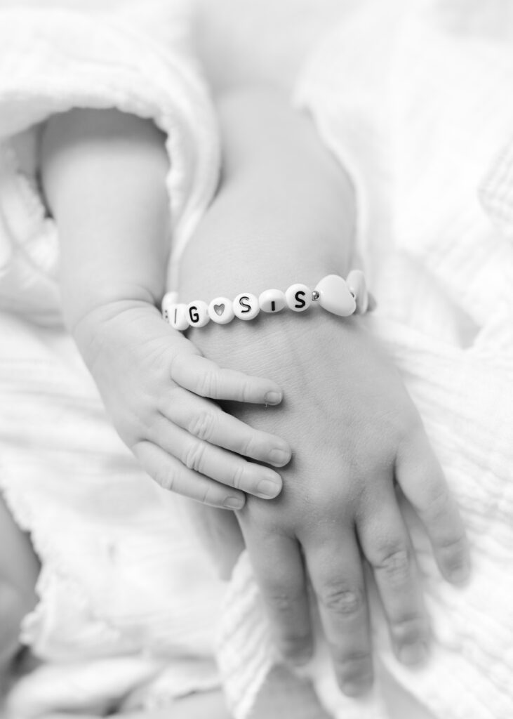 Close up black and white photo of a toddler and baby's hand with a bracelet that says "big sis" Photograph by Maegan R Photography. 