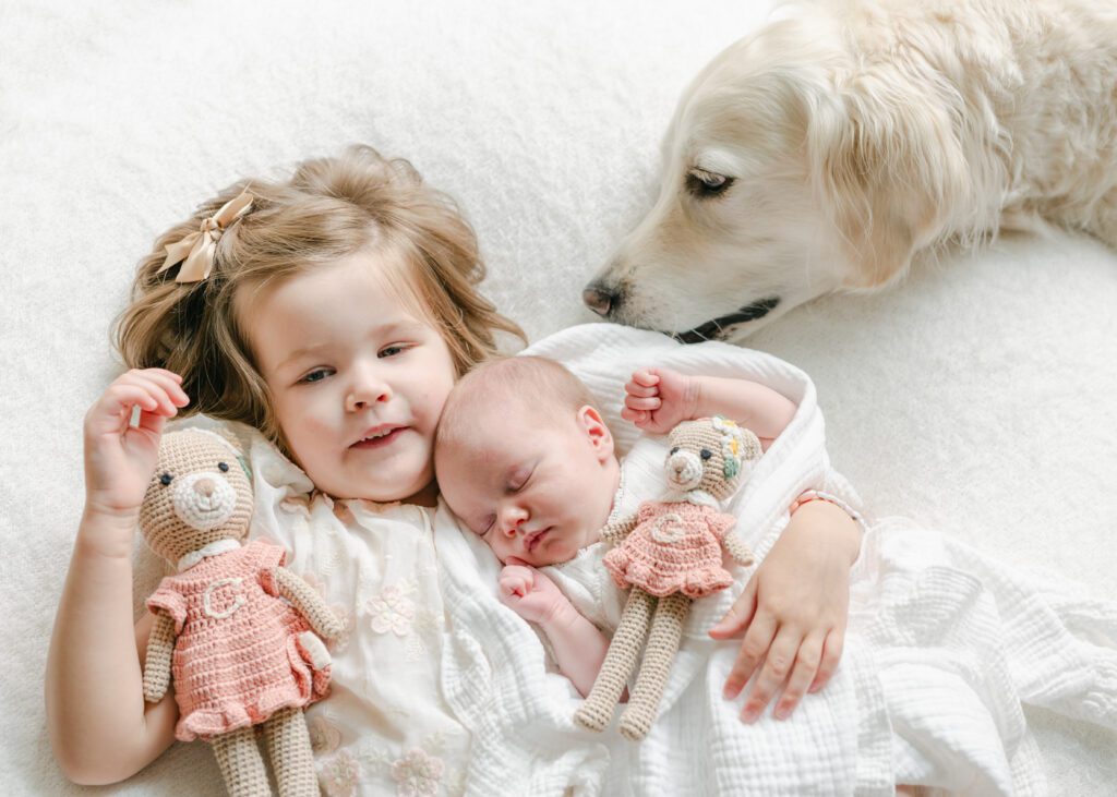 photo of a toddler, newborn baby and white dog snuggling on a white blanket. Photograph by Maegan R Photography. 