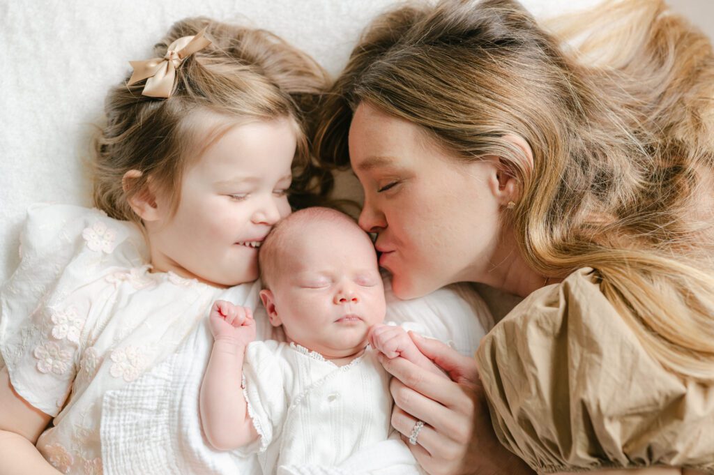 Photo of a mom with her toddler and newborn daughters.  The mom and daughter are kissing the baby, laying on a white blanket. Photograph by Maegan R Photography. 