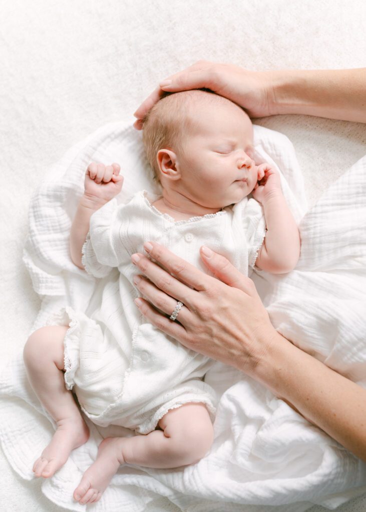 Photo of a baby sleeping on a white blanket with the mothers hand cradling her head.  Photograph by Maegan R Photography. 