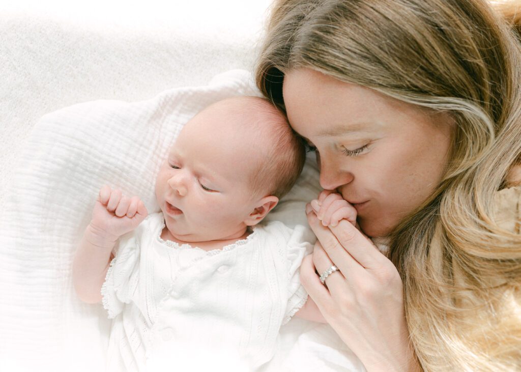 Close up photo of a mom kissing her newborn baby's hand as they lay on a white blanket. Photograph by Maegan R Photography. 